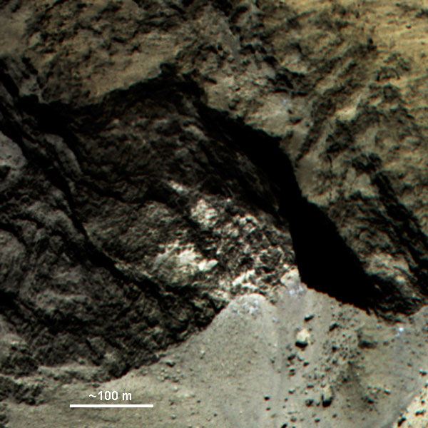 Das Bild zeigt Farbkomposit eines Teils von 67P. Die felsige Oberfläche enthält auffällige helle Flecken. Am Boden unterhalb der Felswand ist zudem Material sichtbar, das heller und blauer ist als die Umgebung. Dabei könnte es sich um Blöcke (blauen) Wassereises handeln.